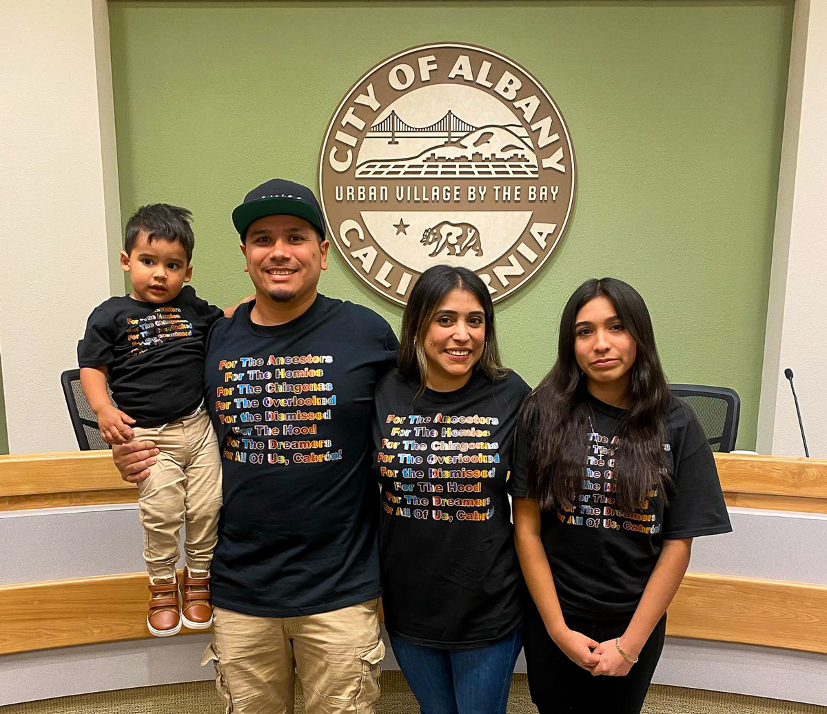 A photo of four people standing together in city council chambers. Behind them is the seal of the City of Albany.