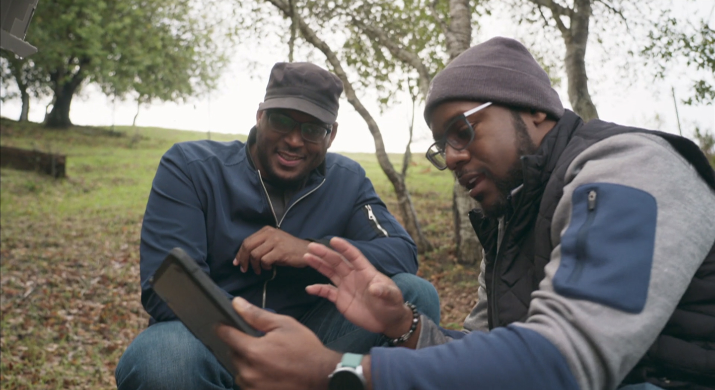 A photo of two men crouching in a park to look at a small tablet