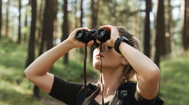 A woman looks through binoculars in a wooded environment.