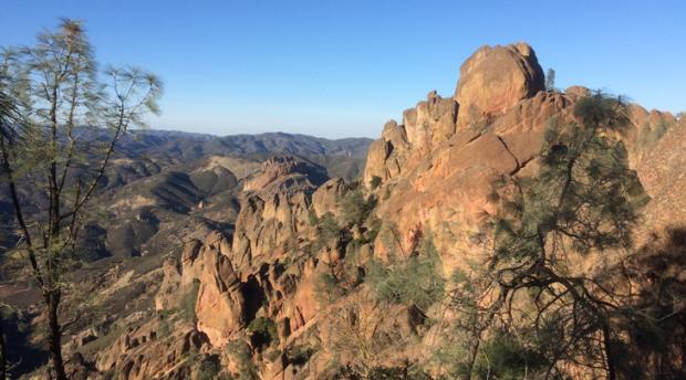 A photo of Pinnacles National Park, showing a mountain range, green trees, and the blue sky.