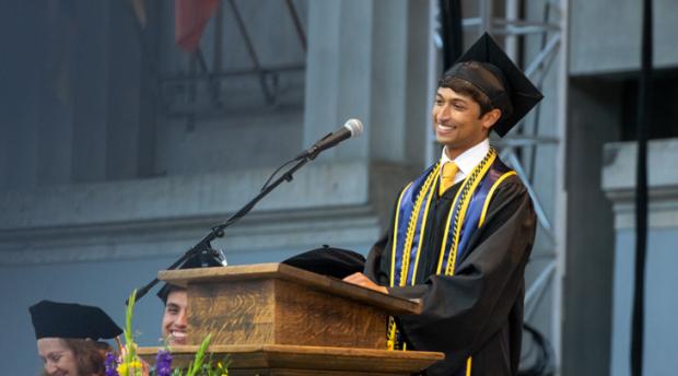 A photo of a man wearing graduation regalia smiling from behind a lectern.