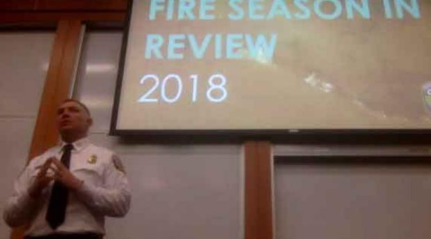 A man stands in front of a presenting screen that reads "fire season in review 2018"