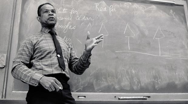 A black and white photo of a man standing in front of a chalkboard in a classroom environment.